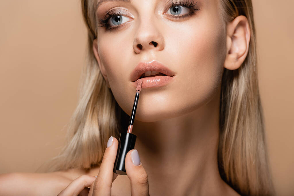 νεαρή γυναίκα με τέλειο πρόσωπο εφαρμογή παστέλ ροζ lip gloss απομονώνονται σε μπεζ - Φωτογραφία, εικόνα