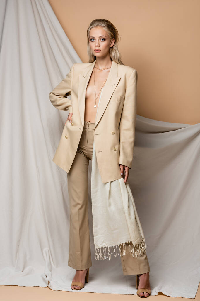 vue pleine longueur de la femme en costume élégant posant avec la main sur la hanche près du tissu sur fond beige - Photo, image