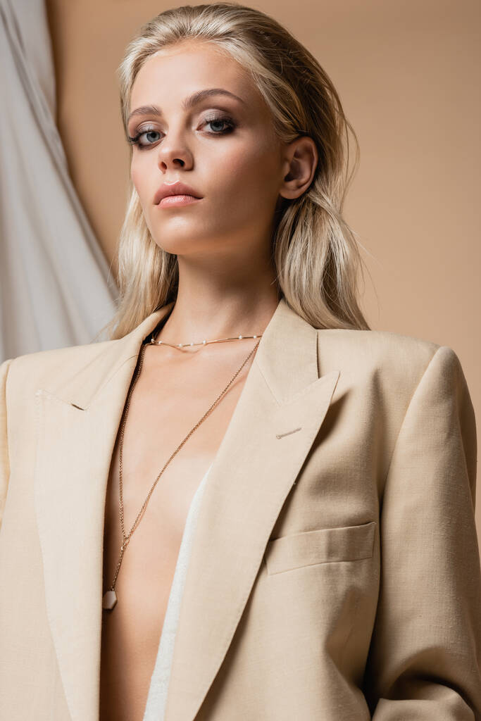 low angle view of blonde woman in unbuttoned blazer and golden necklace on beige draped background - Photo, Image