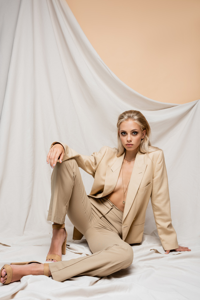 sensual woman in trendy suit looking at camera while sitting on beige background with drapery - Photo, Image