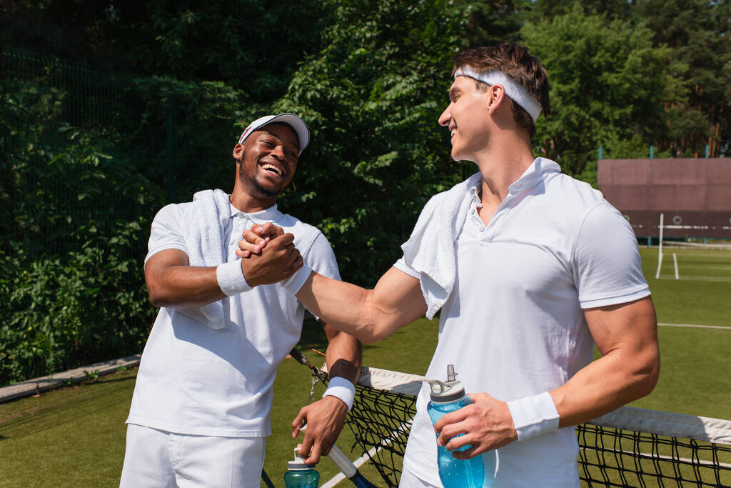 Lächelnde multiethnische Tennisspieler mit Sportflaschen schütteln Hände auf dem Platz  - Foto, Bild