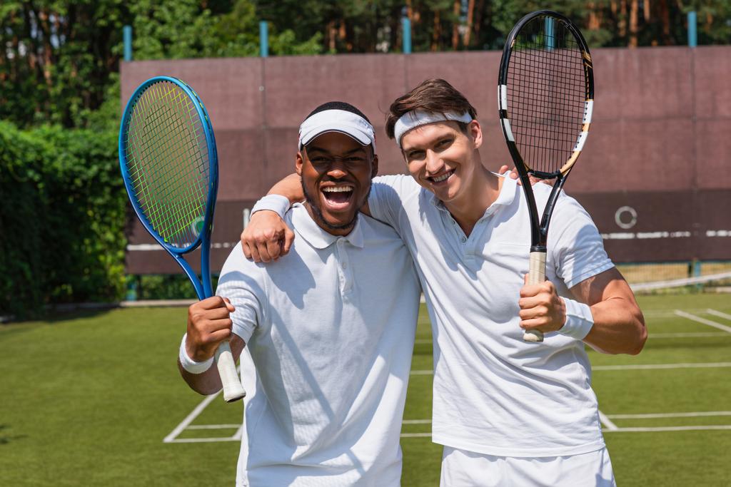 Cheerful multiethnic tennis players hugging on court  - Photo, Image