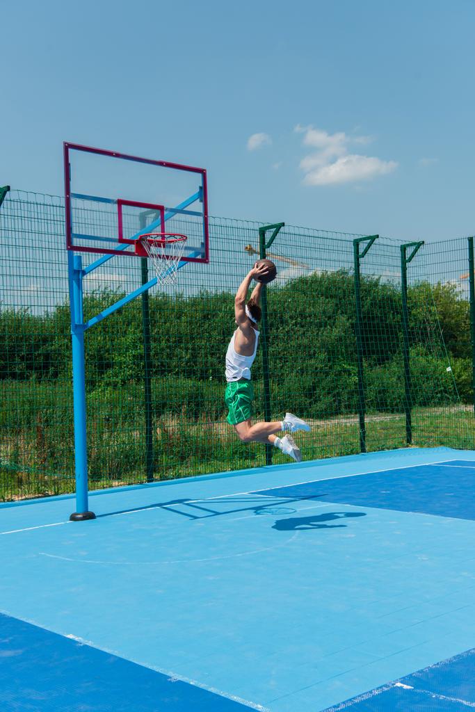 Jovem desportista com bola de basquete pulando sob aro no playground  - Foto, Imagem