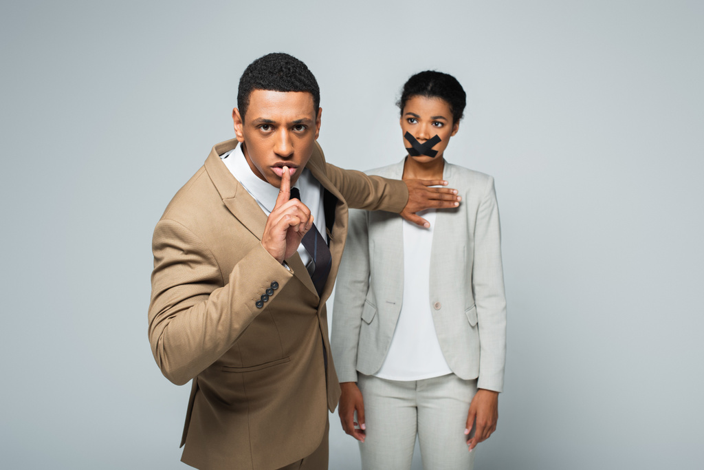 arrogante Afro-Amerikaanse zakenman toont zwijgteken en wegduwen zakenvrouw met scotch tape op de mond geïsoleerd op grijs - Foto, afbeelding