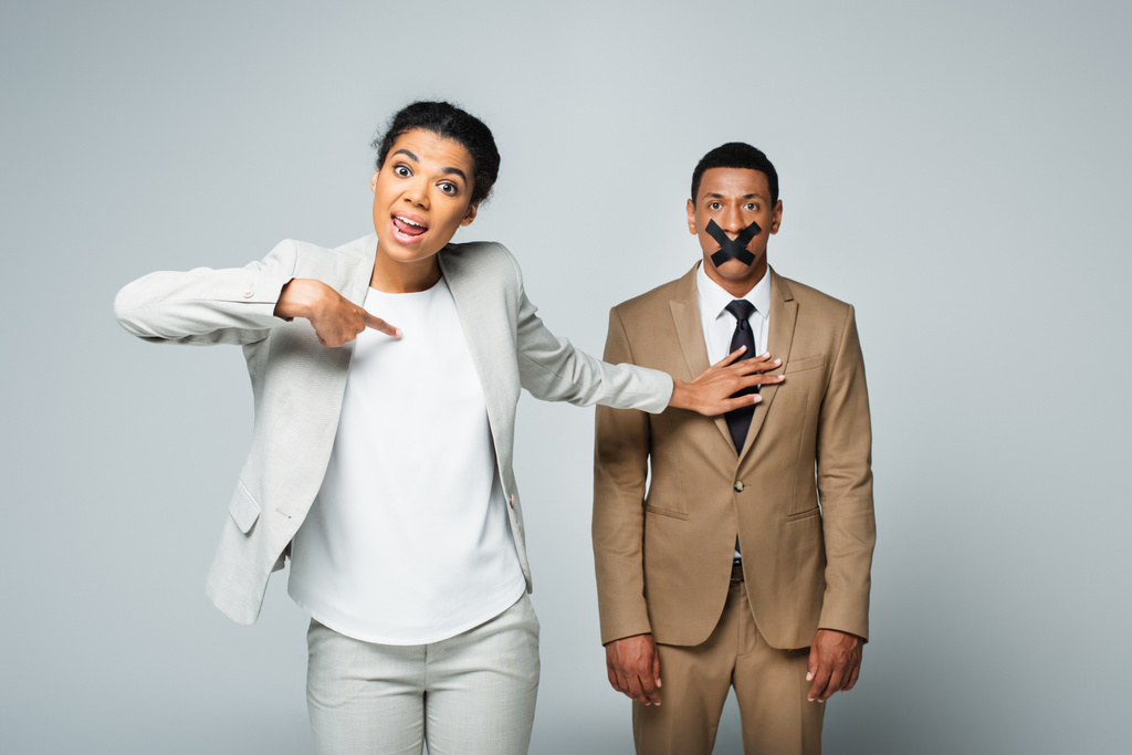 arrogante afrikanisch-amerikanische Geschäftsfrau zeigt auf sich selbst und stößt Manager mit Klebeband auf dem Mund isoliert auf grau - Foto, Bild
