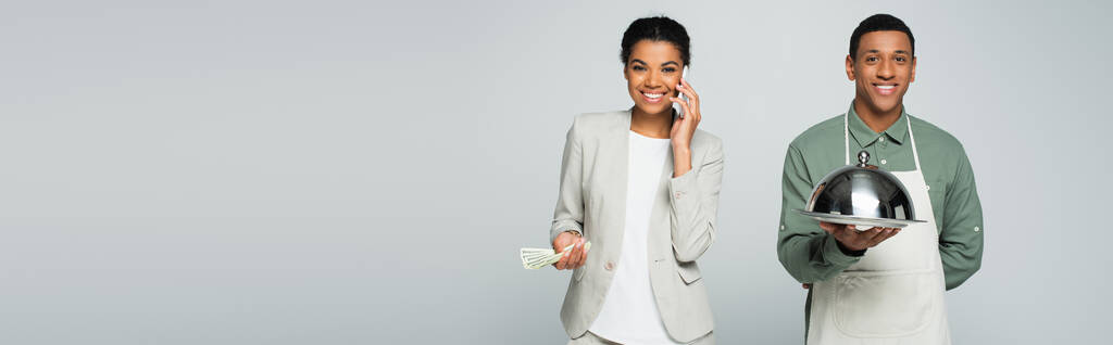 feliz mujer de negocios afroamericana sosteniendo billetes de dólar y hablando en un teléfono inteligente cerca de camarero aislado en gris, bandera - Foto, imagen
