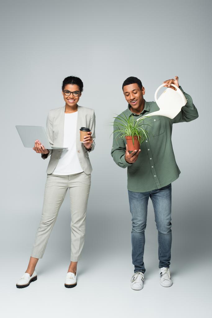 boldog afro-amerikai üzletasszony kezében kávé menni, és laptop mellett kertész öntözés növény szürke - Fotó, kép