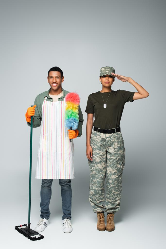 comprimento total do soldado fêmea na tampa perto do limpador masculino no avental com esfregão e escova de poeira no cinza  - Foto, Imagem