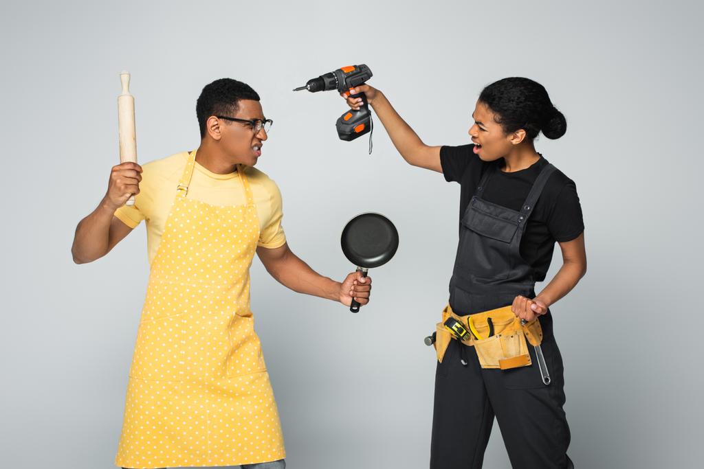 angry african american man in apron holding rolling pin and frying pan while fighting with repairwoman isolated on grey - Photo, Image