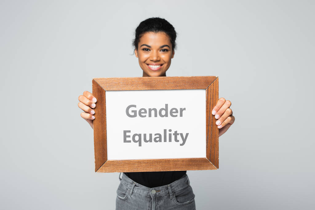 mujer afroamericana sonriente sosteniendo pancarta con letras de igualdad de género aisladas en gris - Foto, Imagen