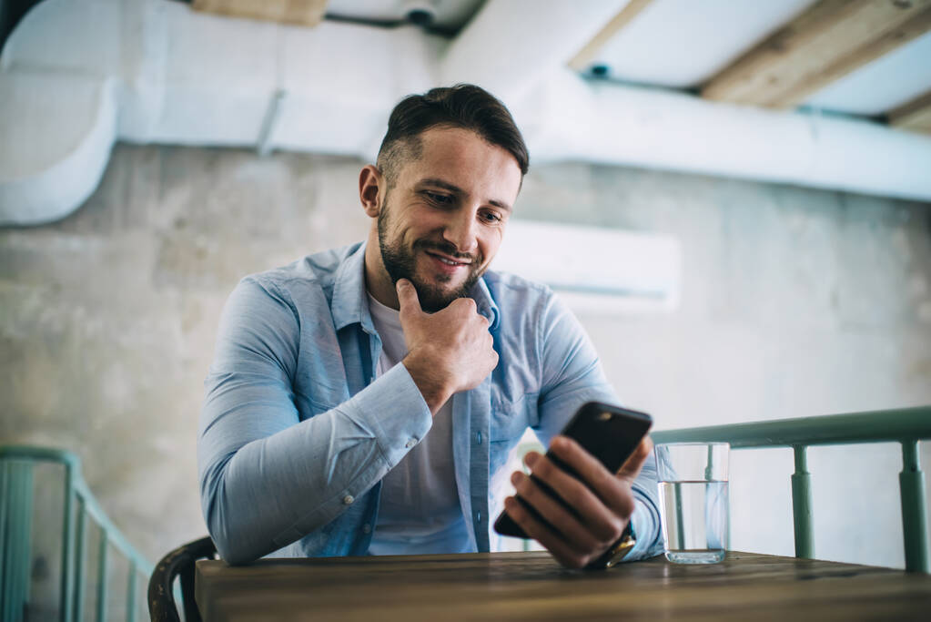 Pensive hipster kaveri lukee viestin matkapuhelimella chattailuun sosiaalisissa verkostoissa tauolla kahvilassa sisustus, hymyilevä uros tuhatvuotinen katsella video älypuhelimella nauttia sovellus ja 4G-yhteys - Valokuva, kuva