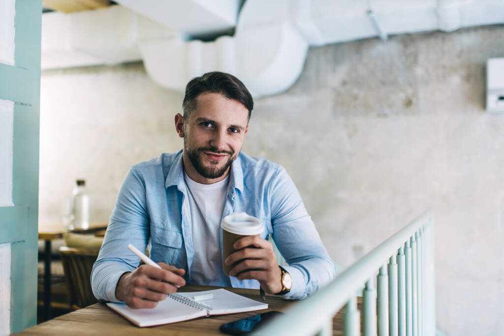 Portret wesołego białego pisarza wykonującego odległą pracę w kawiarni trzymającego długopis do robienia notatek, uśmiechniętego hipstera cieszącego się kawą i uczącego się do szkoły biznesu patrzącego przed kamerą - Zdjęcie, obraz