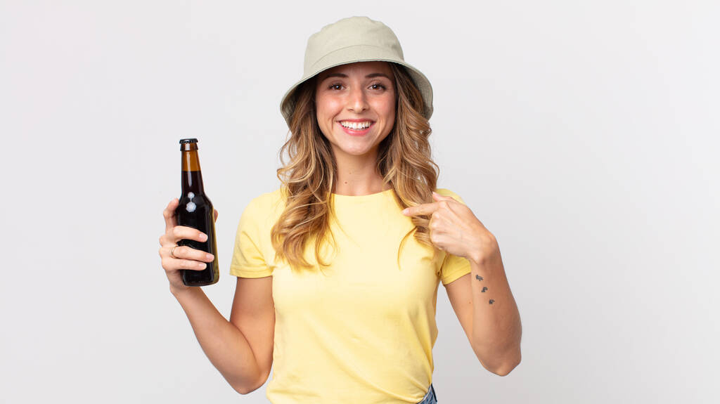 mujer bastante delgada sintiéndose feliz y señalándose a sí misma con una excitada y sosteniendo una cerveza. concepto de verano - Foto, imagen