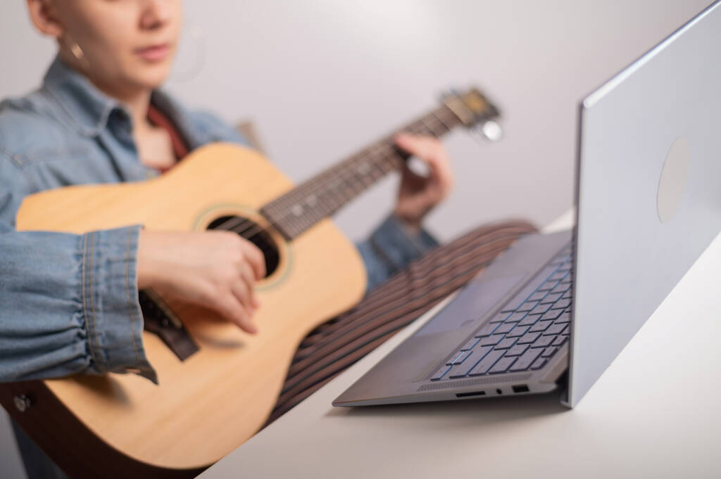 Junge kaukasische Frau mit kurzen blonden Haaren spielt Gitarre und schaut sich Trainingsvideo auf Laptop an. - Foto, Bild