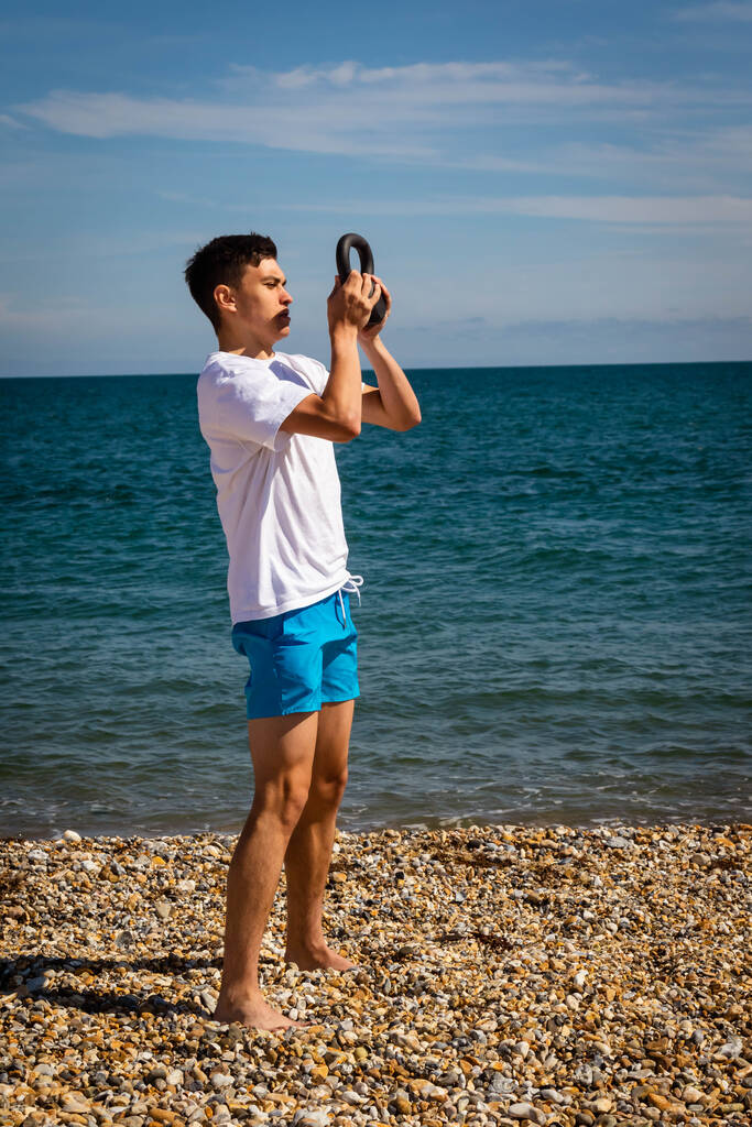 Un adolescente caucásico de 18 años sin camisa en una playa haciendo ejercicio con un peso de pesas usando ambas manos - Foto, Imagen