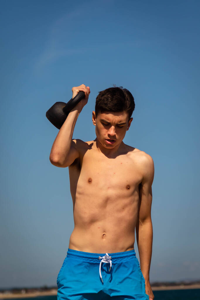 Un adolescent blanc torse nu de 18 ans en maillot de bain faisant de l'exercice avec un poids de kettlebell - Photo, image