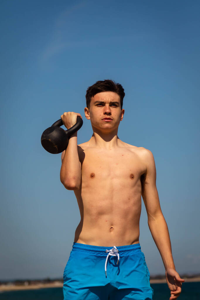 Un ragazzo caucasico di 18 anni senza maglietta adolescente in costume da bagno che si esercita con un peso kettlebell - Foto, immagini