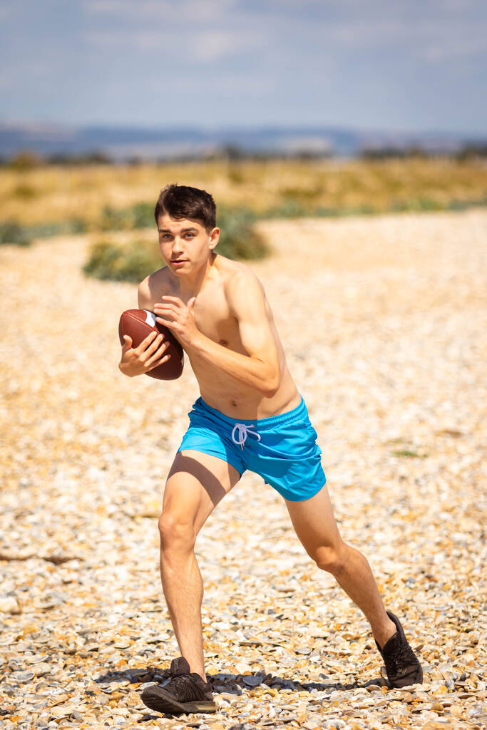 18-летний кавказский юноша без рубашки бегает по пляжу с американским футболом - Фото, изображение