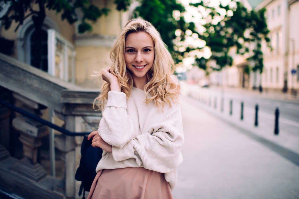 Femme blonde joyeuse en tenue décontractée souriant et regardant la caméra tout en se tenant debout sur une vieille rue historique et en s'appuyant sur une balustrade en pierre - Photo, image