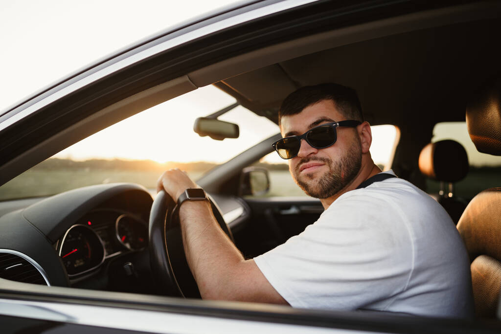 Vousatý muž řidič v slunečních brýlích sedí v autě a dívá se do kamery - Fotografie, Obrázek