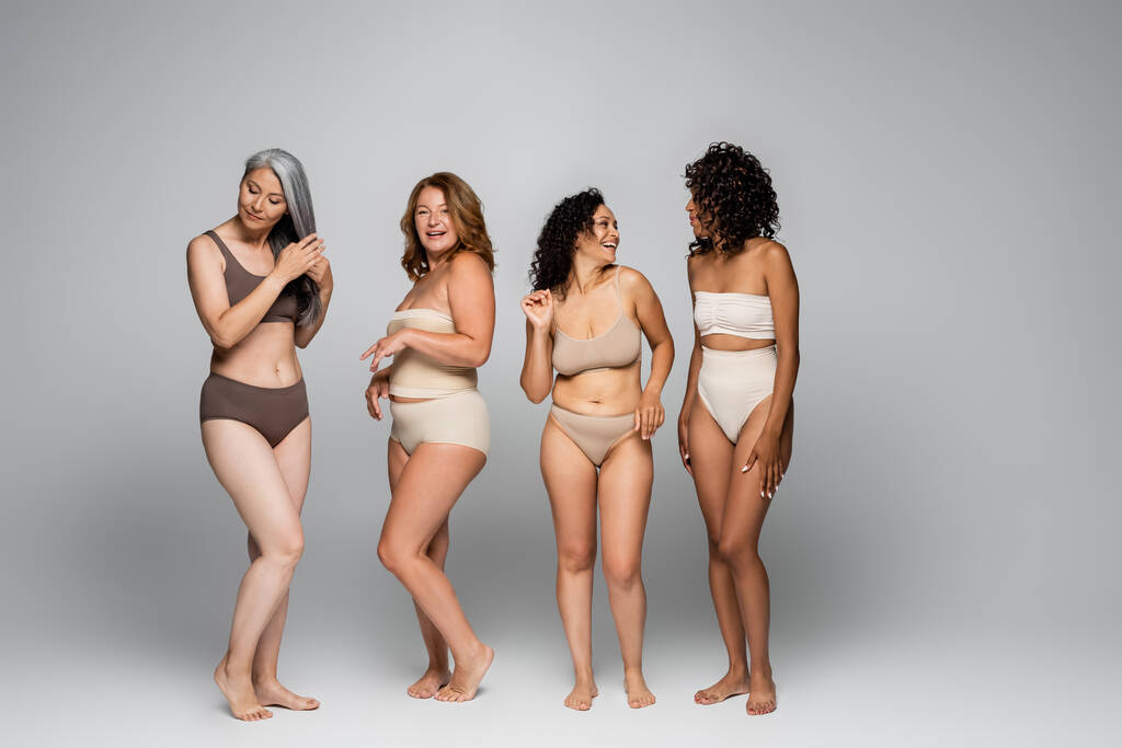 Χαμογελώντας διαφυλετικές γυναίκες σε εσώρουχα στέκεται σε γκρι φόντο, το σώμα θετική έννοια - Φωτογραφία, εικόνα