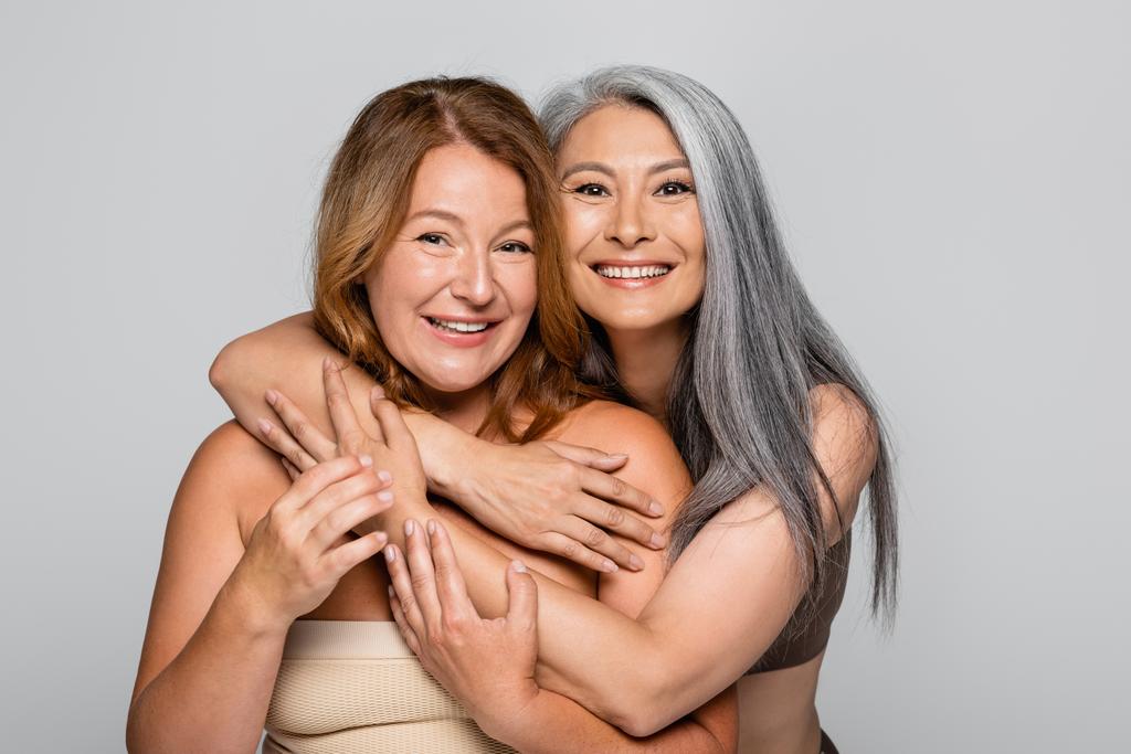 Smiling asian woman hugging friend in bra isolated on grey, body positive concept - Photo, Image