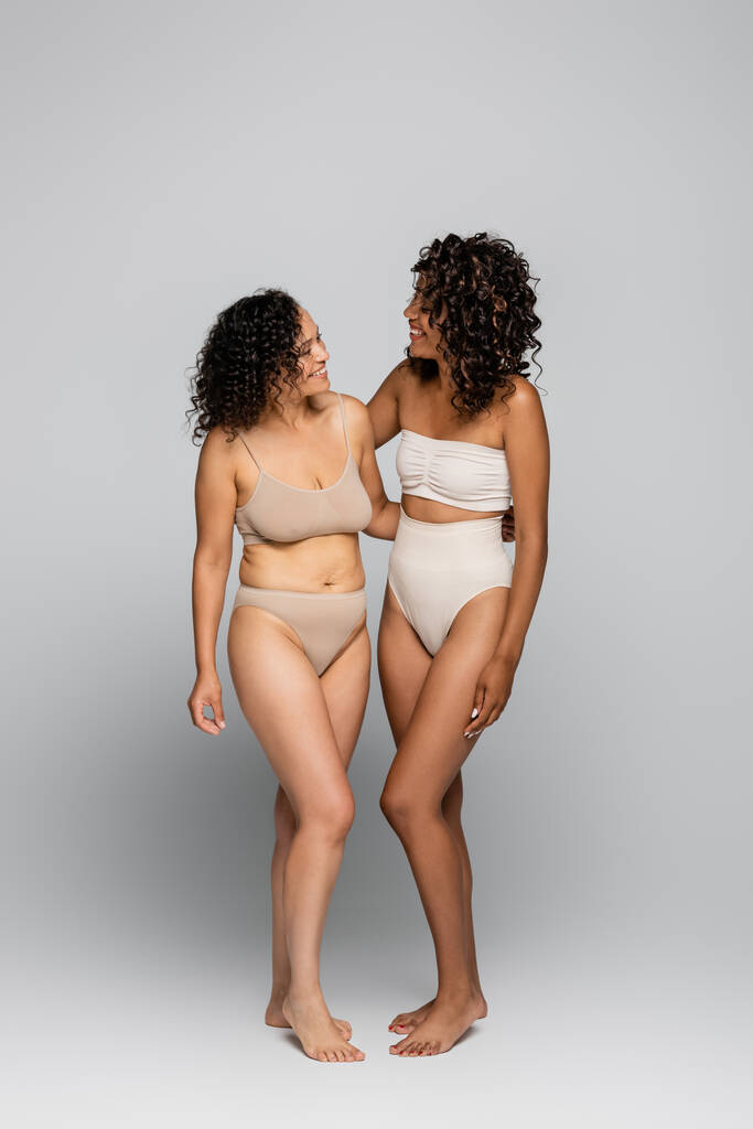 Gülümseyen Afro-Amerikan kadınları iç çamaşırlarıyla gri arka planda sarılıyorlar, vücut pozitifliği - Fotoğraf, Görsel