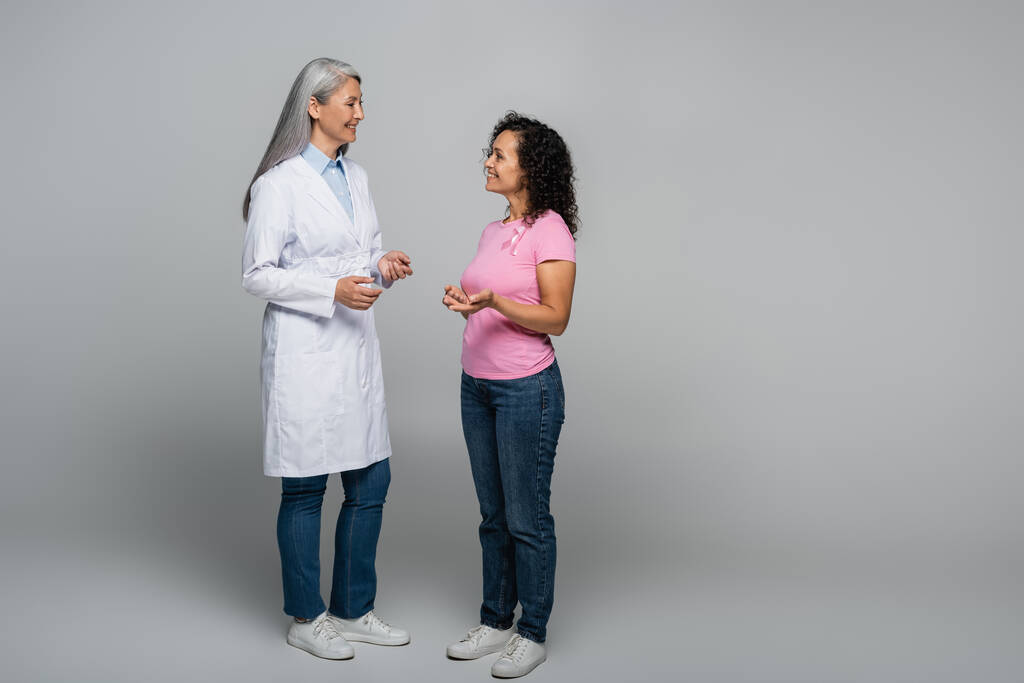 Allegro medico asiatico che parla con il paziente afro-americano con nastro di consapevolezza del cancro al seno su sfondo grigio  - Foto, immagini