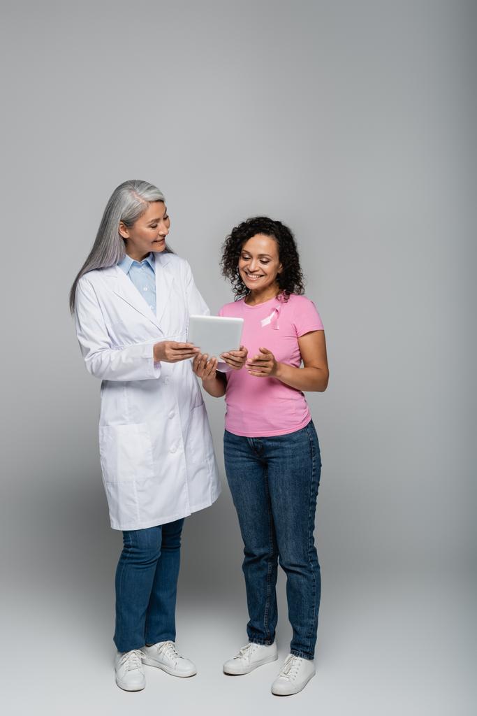 Cheerful asian doctor holding digital tablet near african american patient with pink ribbon on grey background  - Photo, Image