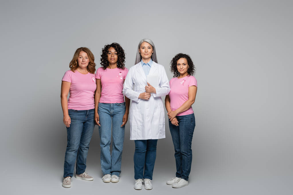Азиатский врач стоит рядом с межрасовыми женщинами с розовыми лентами на сером фоне  - Фото, изображение
