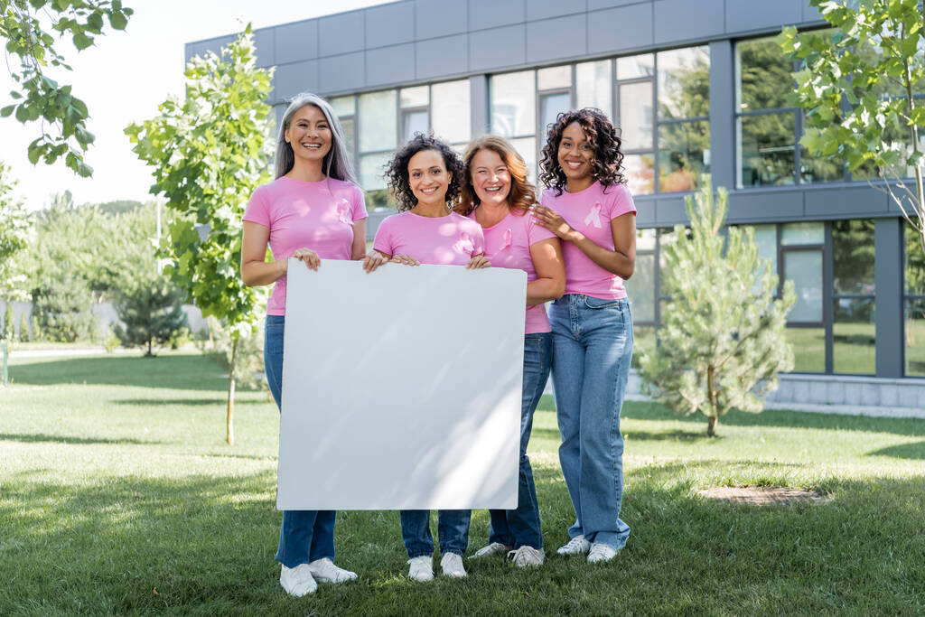 Uśmiechnięte kobiety z różowymi wstążkami trzymające plakietkę na trawniku  - Zdjęcie, obraz