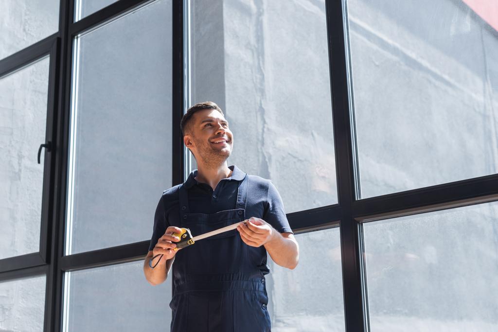 happy workman standing near large windows with measuring tape - Photo, Image