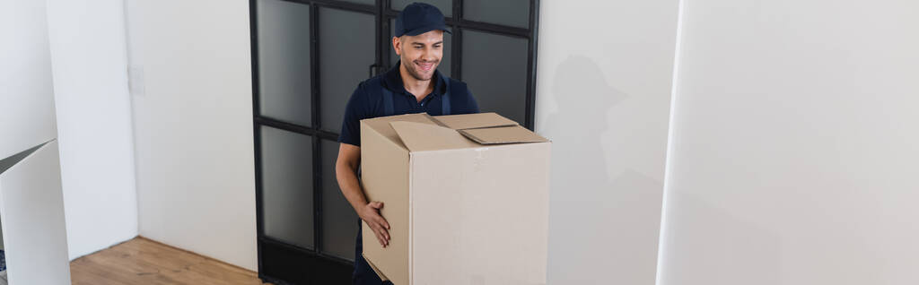 joyful mover in overalls carrying large carton box in apartment, banner - Photo, Image