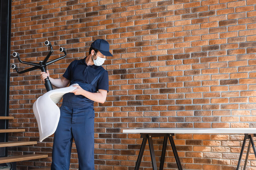 travailleur dans le masque de protection tenant chaise de bureau près du mur de briques et table - Photo, image