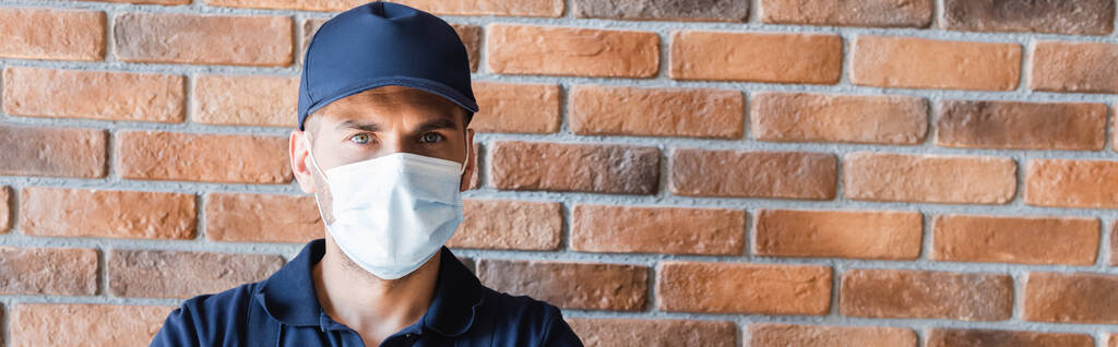 workman in cap and medical mask looking at camera near brick wall, banner - Photo, Image