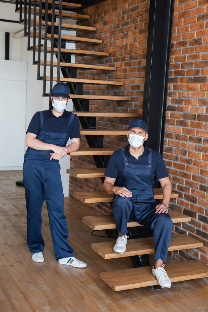 bricoleurs en uniforme et masques médicaux regardant caméra près des escaliers et mur de briques - Photo, image
