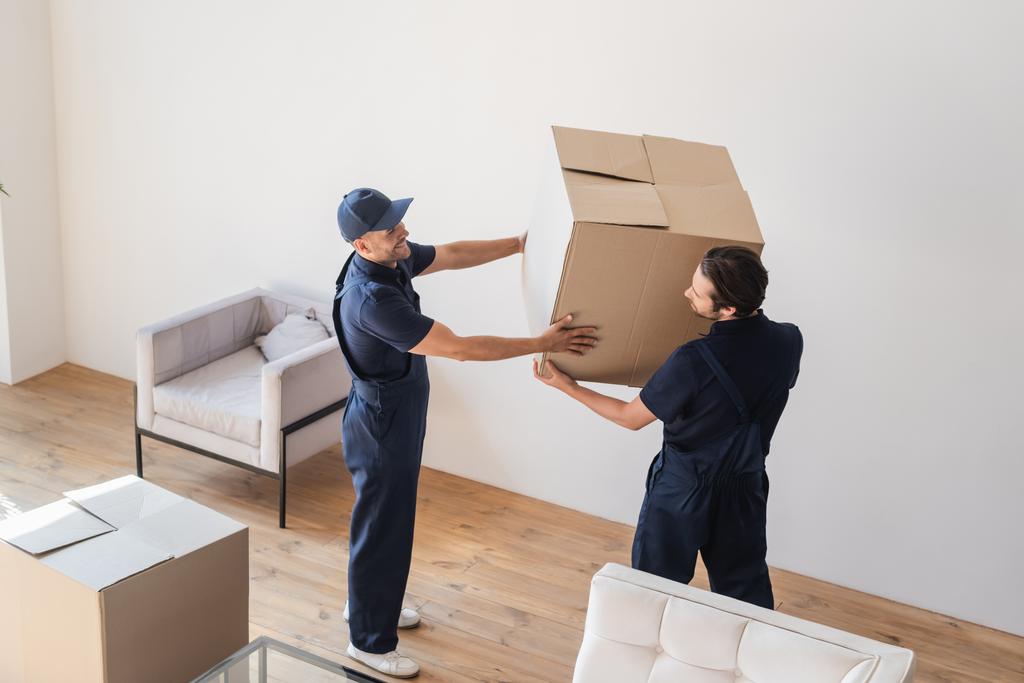 klusjesmannen in uniform die een kartonnen doos in de woonkamer dragen - Foto, afbeelding