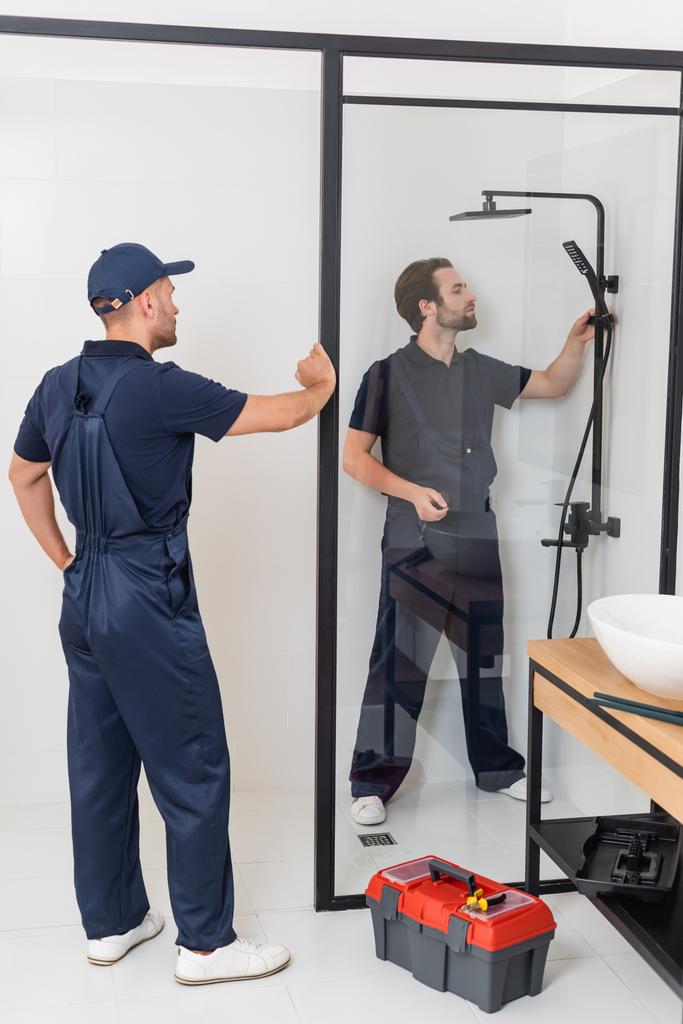 werknemer staan met de hand op heup in de buurt loodgieter controleren douche in de badkamer - Foto, afbeelding