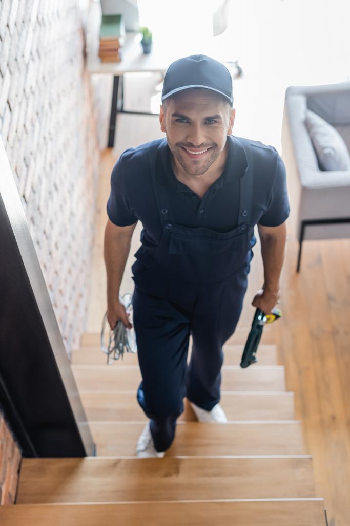vue grand angle de l'ouvrier souriant marchant à l'étage avec des fils et des outils - Photo, image