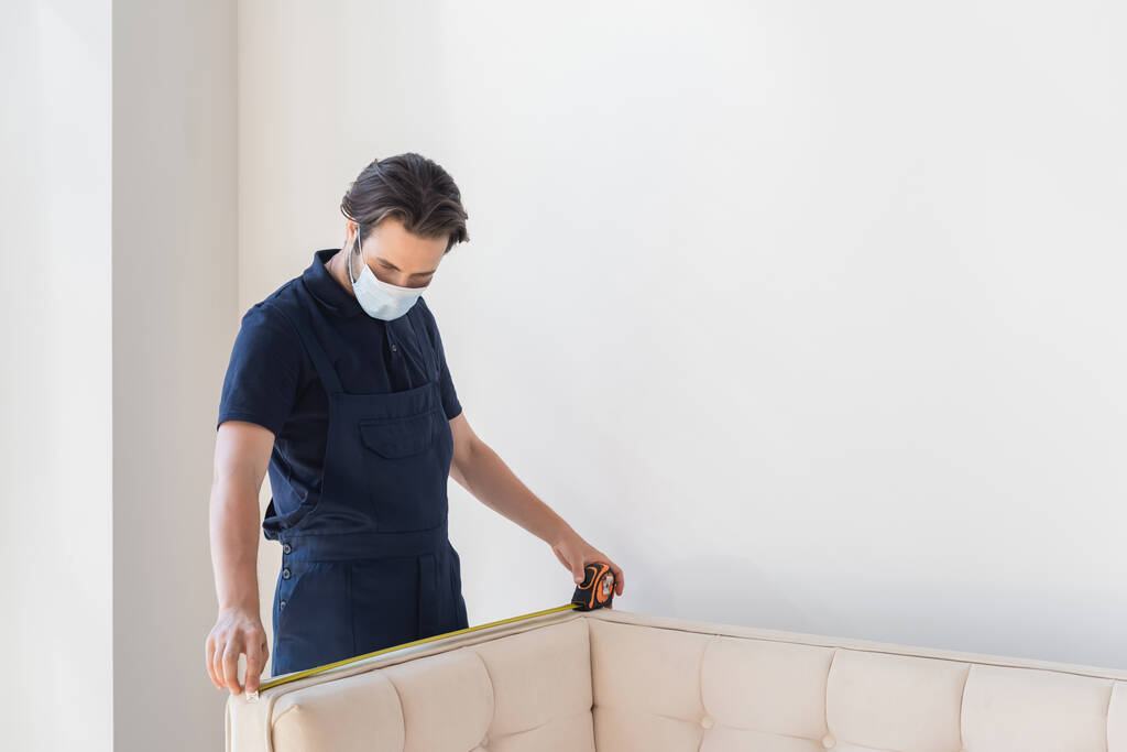 pracovník v lékařské masce měřící gauč u bílé stěny - Fotografie, Obrázek