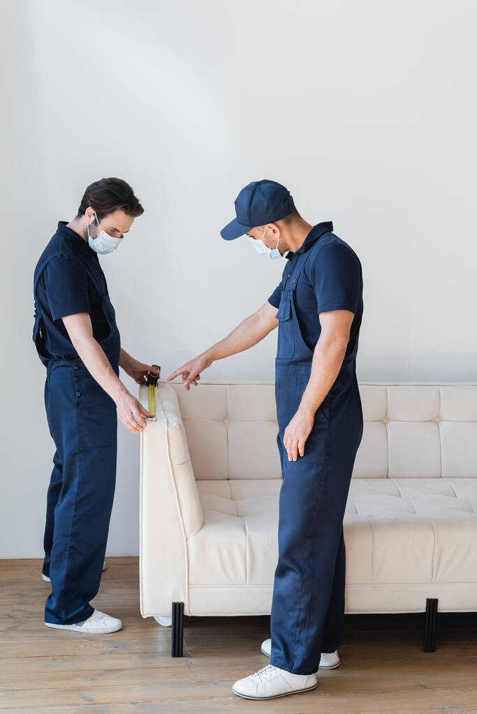 ouvrier en masque de sécurité pointant vers un collègue mesurant un canapé blanc - Photo, image