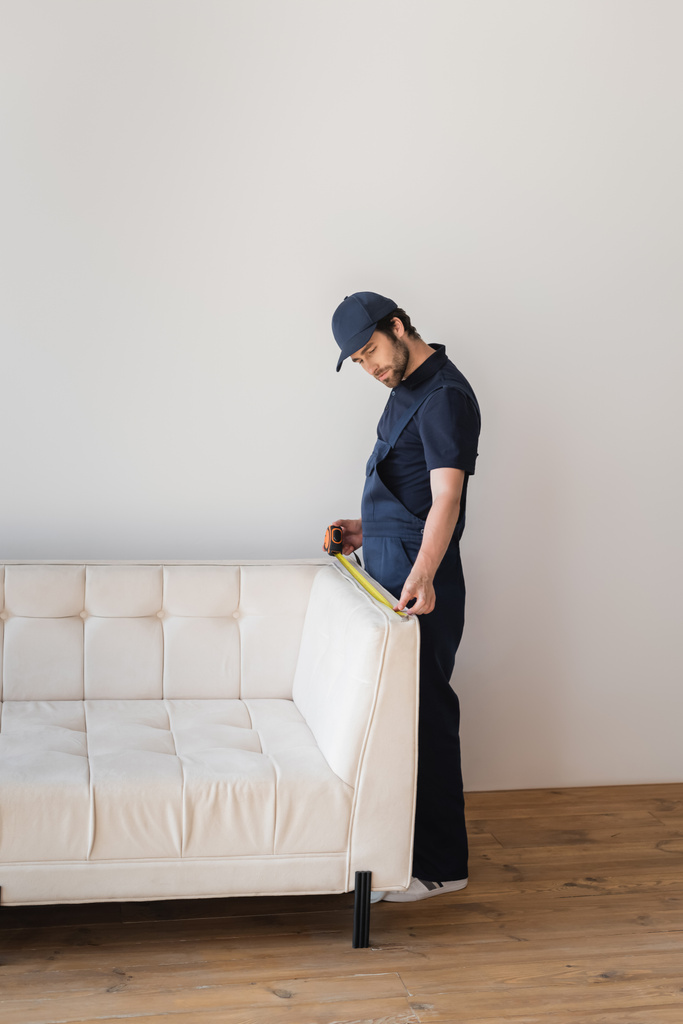 рабочий в форме, меряющий белый диван в гостиной - Фото, изображение