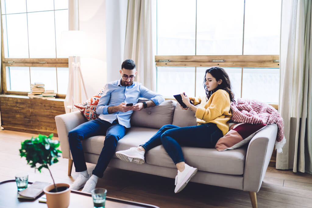 Пара зайнятих віддалених працівників, які переглядають сенсорний екран телефону і цифрового планшета, лежачи на дивані в затишній світлій вітальні вдома
 - Фото, зображення
