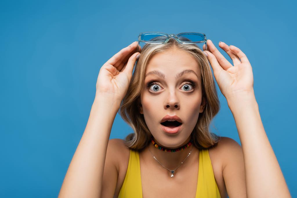 σοκαρισμένη νεαρή γυναίκα προσαρμόζοντας μοντέρνα γυαλιά ηλίου και κοιτάζοντας κάμερα απομονωμένη στο μπλε - Φωτογραφία, εικόνα