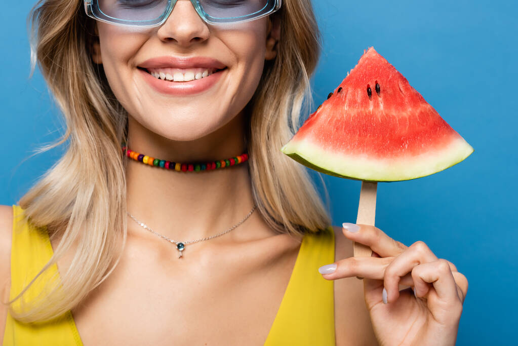 bijgesneden mening van gelukkige jonge vrouw in zonnebril houden lolly stick met watermeloen geïsoleerd op blauw - Foto, afbeelding