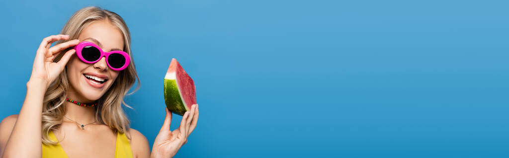 joyful young woman adjusting pink sunglasses and holding slice of watermelon isolated on blue, banner - Photo, Image