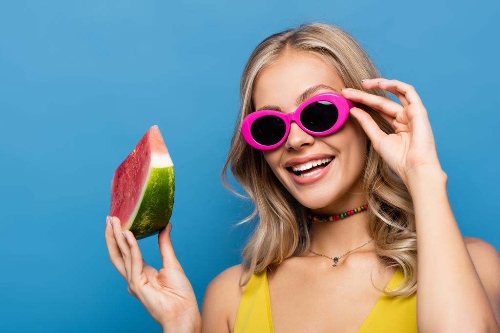 радісна молода жінка регулює рожеві сонцезахисні окуляри і тримає шматочок кавуна ізольовано на синьому
 - Фото, зображення