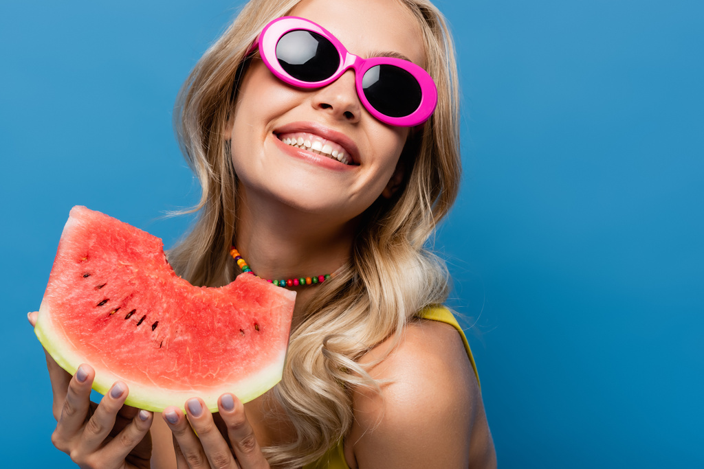 весела молода жінка в рожевих сонцезахисних окулярах тримає солодкий кавун ізольовано на синьому
 - Фото, зображення