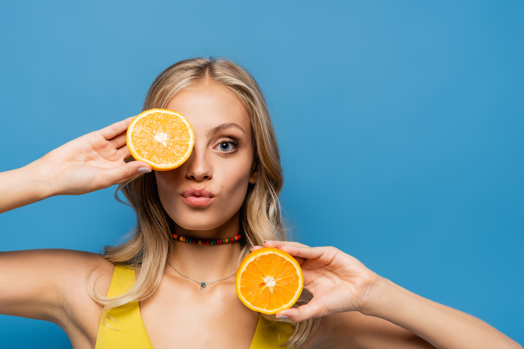 блондинка молодая женщина надувая губы и закрывая глаза оранжевым наполовину изолированы на синий - Фото, изображение