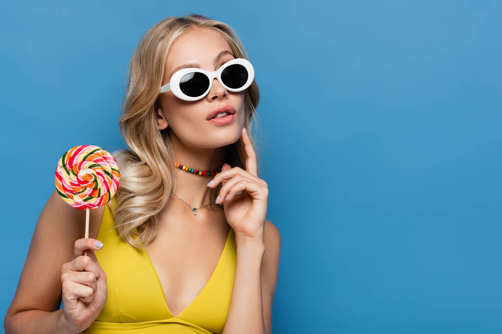 zamyślona młoda kobieta w żółtym bikini i okularach przeciwsłonecznych trzymająca słodki lizak odizolowany na niebiesko - Zdjęcie, obraz